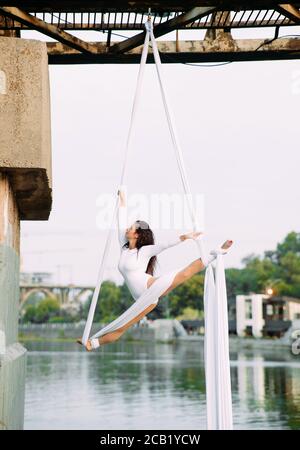 Woman aerialist performs gymnastic split on hanging aerial silk attached to the bridge support against background of sky. Stock Photo