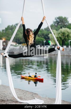 Woman aerialist performs gymnastic split on hanging aerial silk against background of river, floating boat and sky. Stock Photo
