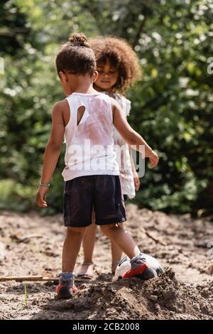 Selective focus of poor african american children in messy clothes playing football on dirty road Stock Photo