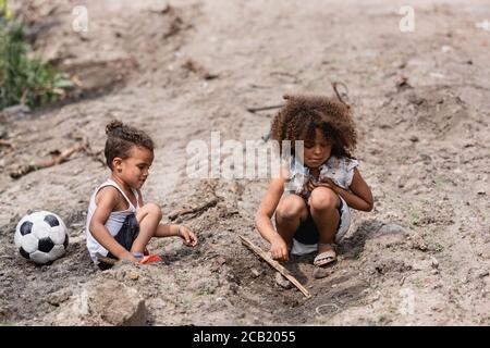 Poor african american kids playing on dirty road near football in slum Stock Photo