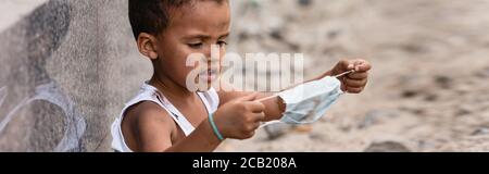 panoramic shot of poor african american kid holding dirty medical mask outside Stock Photo