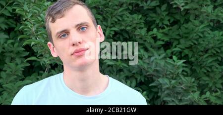 Young blue-eyed man looking tiredly at the camera on a background of green bushes Stock Photo