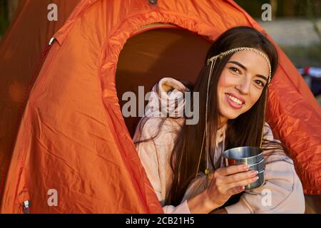 Cheerful female traveler with hot drink sitting in camp tent Stock Photo