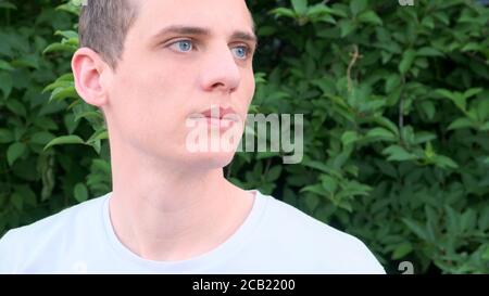 Portrait of a young very handsome man with blue eyes and blond hair looking to the side against a background of greenery Stock Photo