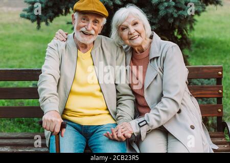 Grey haired couple holding hands and smiling at camera on bench in park Stock Photo