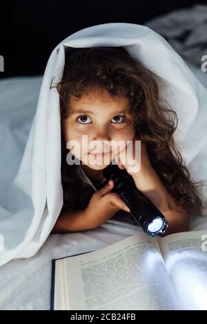 Beautiful little girl reads a book with a flashlight in the bedroom under a blanket