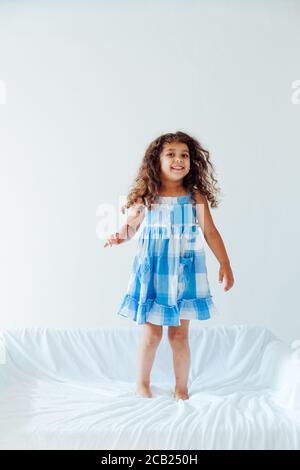 Portrait of a beautiful little curly girl in a blue dress on a white sofa Stock Photo