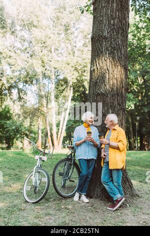 Elderly couple smiling at each other while holding paper cups near tree and bikes in park Stock Photo