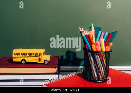 school bus model on staked books near pen holder with stationery on notebook near green chalkboard Stock Photo