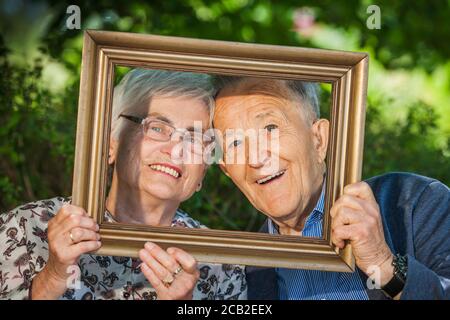 Head-and-shoulder portrait of an 80-year-old retired couple with picture frames in front of their faces, happily posing in front of a blurred green he Stock Photo