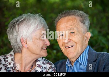 Head-and-shoulder portrait of an 80 year old pensioner couple against blurred green background. Woman looks at man. Man looks to the camera Stock Photo