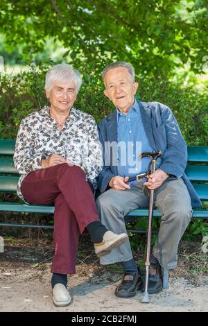 Full body portrait of an 80 year old retired couple on a park bench in the shade of a canopy in summer looking joyfully into the camera. Stock Photo