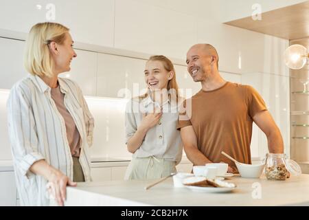 Warm-toned waist up portrait of modern family laughing happily while enjoying breakfast together standing by table in minimal kitchen interior, copy space Stock Photo