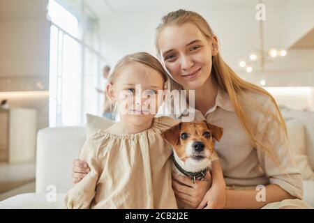 Warm-toned portrait of two sisters posing with pet dog and looking at camera while sitting on couch in minimal home interior, copy space