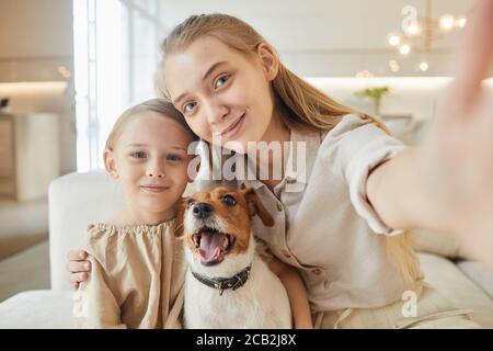 Warm-toned portrait of two sisters taking selfie with pet dog and looking at camera while sitting on couch in minimal home interior, copy space Stock Photo