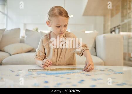Warm-toned portrait of cute little girl solving jigsaw puzzle while enjoying time indoors at home, copy space