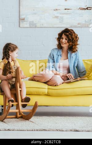 smiling nanny looking at kid riding rocking horse while sitting on sofa with book Stock Photo