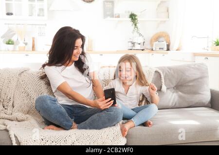 Attractive young mom and little daughter playing online games at home using smartphone, having video chat or watching movie. Distance education. Video Stock Photo