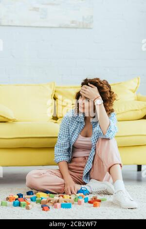 exhausted young woman sitting on floor near multicolored blocks and touching forehead Stock Photo