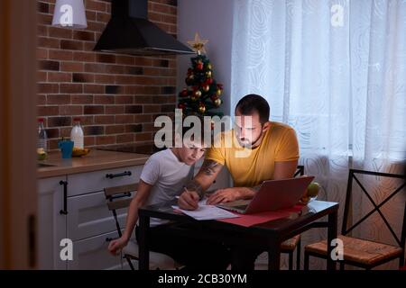 caucasian child boy doing homework at home, sitting in kitchen with father, use modern laptop. studying and school concept Stock Photo