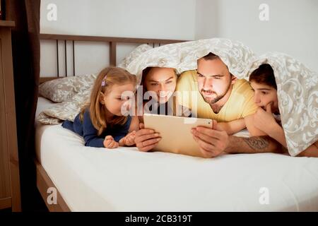 lovely caucasian family spending time together at morning, lying on bed under blanket and watching on screen of tablet, watching film together Stock Photo