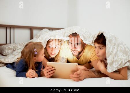 caucasian family, mother father and children lying under blanket on bed watching video on tablet, leisure time at home. Stock Photo