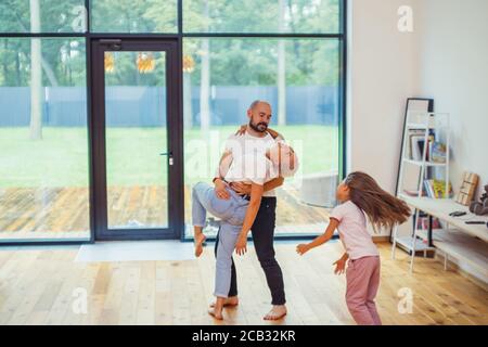 Young caucasian family happy at home. Parents dance with each other while their daughter running. White interior, big panoramic window. Family concept Stock Photo