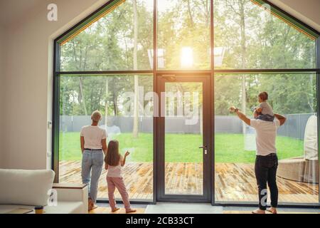 Enjoying family look at window, dream. baby sits on dad's shoulders, daughter stand with mother Stock Photo