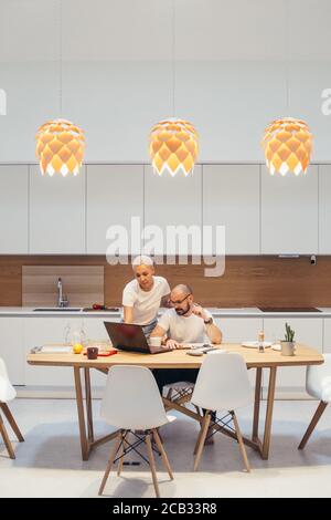 Beautiful european couple in kitchen use laptop. Man is sitting with laptop, woman next to him communicate, discuss together new design for home, new Stock Photo
