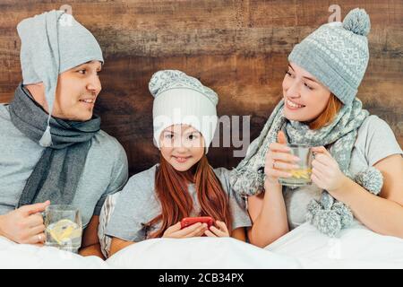 sick family lying on bed together, cute kid girl lying between mother and father and hold smartphone in hands Stock Photo