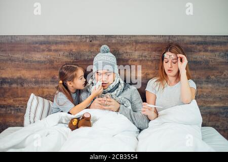 sick caucasian man lying on bed with high temperature with careful wife and kid girl, wearing hat and scarf, isolated in bedroom Stock Photo