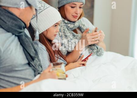 ill family lying on bed together, beautiful kid girl lying between mother and father and hold smartphone in hands Stock Photo