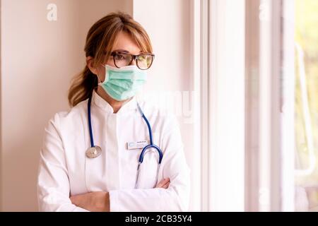Female doctor wearing face mask and looking careworn while looking out the window during coronavirus spreading rapidly in the country. Stock Photo