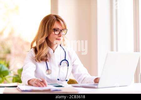 Portrait shot of middle aged female doctor sitting at desk and working on laptop in doctor’s office. Stock Photo