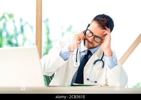 Shot of stressed male doctor sitting at desk behind his laptop and making a call in the doctor’s room. Stock Photo