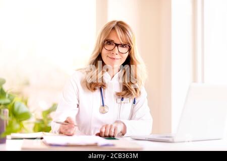 Portrait shot of smiling female doctor sitting at desk while writing something and working on laptop in doctor’s office. Stock Photo
