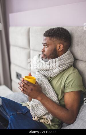 Young African businessman being upset and distempered with his illness, wrapped in knitted thick warm scarf lying on bed while holding a mug with hot Stock Photo