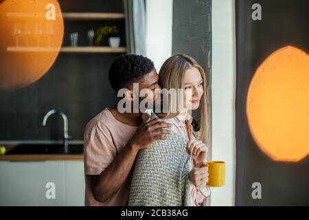 Handsome african guy kissing his caucasian girlfriend on the neck from backside, standing at home in the kitchen room with modern interior Stock Photo