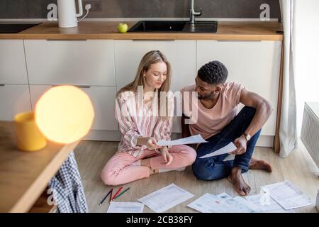 Young mixed-race couple of diverse students in casual wear, sitting together on kitchen floor at home working at their coursework having concentrated Stock Photo