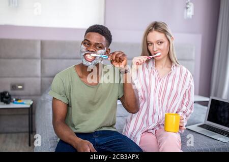 Attractive multiethnic couple brushing their teeth in the morning at home, african man having shaving cream on his cheeks Stock Photo
