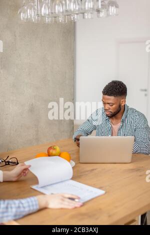 Self-employed young dark-skinned bearded afro-american man working on the laptop while sitting at the table in living room Stock Photo