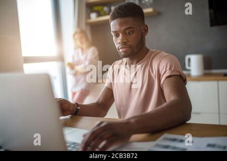 Self-employed young dark-skinned bearded afro-american man working on the laptop while sitting at the table in living room Stock Photo