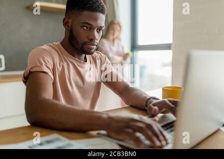 Handsome dark-skinned man having thoughtful and serious facial expression while reviewing finances and paying on-line bills, sitting at desk with docu Stock Photo