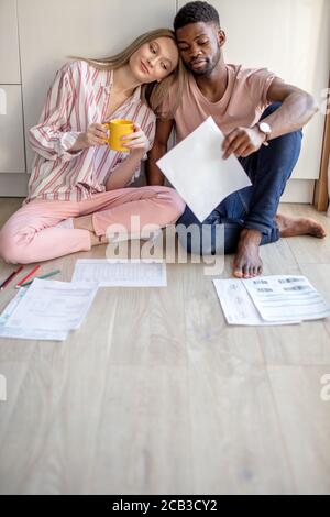 Young mixed-race couple of diverse students in casual wear, sitting together on kitchen floor at home working at their coursework having concentrated Stock Photo