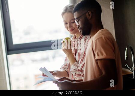 Young mixed-race couple of diverse students in casual wear, standing together next to window reading coursework. Clever multiracial friends studying t Stock Photo