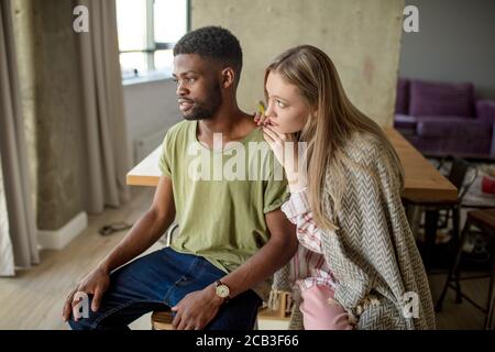 Caucasian blonde woman consoles and supports her african hipster boyfriend, who is having gloomy and frustrated look, couple is sitting in cozy living Stock Photo