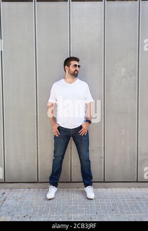 Bearded guy in sunglasses leaning on metallic wall with hands on pocket while looking away Stock Photo