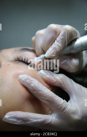 woman lying on couch at beauty salon with closed eyes and enjoying when cosmetologist making eyeliner and eyebrow permanent make up. Professional wear Stock Photo