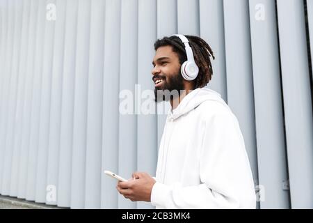 Image of a handsome pleased smiling young man walking outdoors while listening music by headphones and using mobile phone