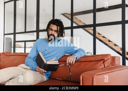 Handsome young african man sitting on a leather couch at home, reading a book Stock Photo